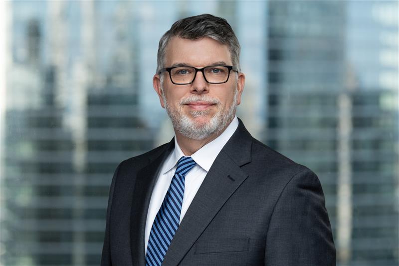 Minsky, McCormick & Hallagan, P.C. Excited to Expand its Federal Litigation Practice by Adding Robert Carpenter & Patricia Capt A man with gray hair, beard, and glasses wearing a dark suit, white shirt, and blue striped tie stands in front of a window with a blurred cityscape background.