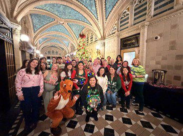 Happy Holidays from Minsky, McCormick & Hallagan, P.C. A group of people in festive sweaters poses in front of a decorated Christmas tree inside a grand hall with arched ceilings; one person is dressed in a reindeer costume.